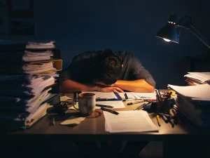 Uitgeputte professional hangend over een rommelig bureau 's nachts, omringd door stapels mappen en papieren, verlicht door een warme bureaulamp.