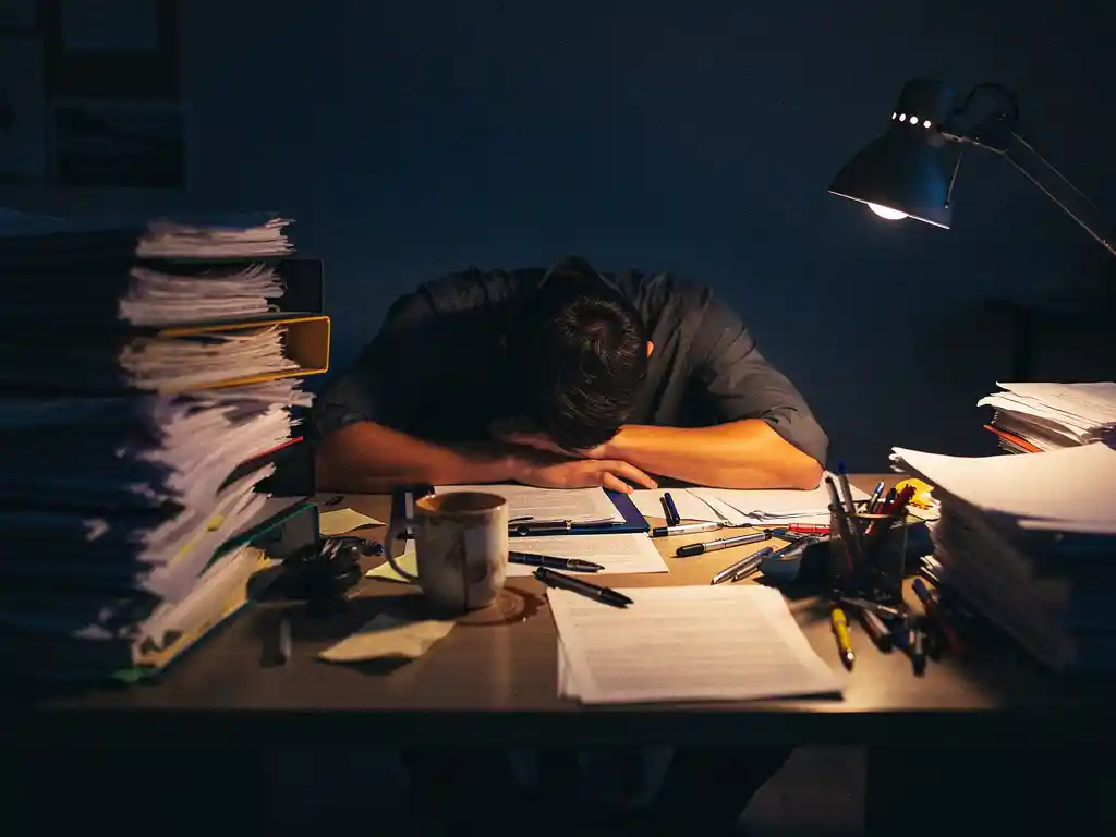 Uitgeputte professional hangend over een rommelig bureau 's nachts, omringd door stapels mappen en papieren, verlicht door een warme bureaulamp.